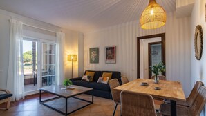 A modern living room with a sofa, dining table, and chairs. There is a large window with curtains, a hanging light fixture, and artwork on the wall. - Los Gavilanes - Isla Canela Beach ICLHG62SP (Isla Canela)