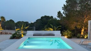 Una piscina rectangular con plataforma blanca, rodeada de plantas y árboles tropicales.