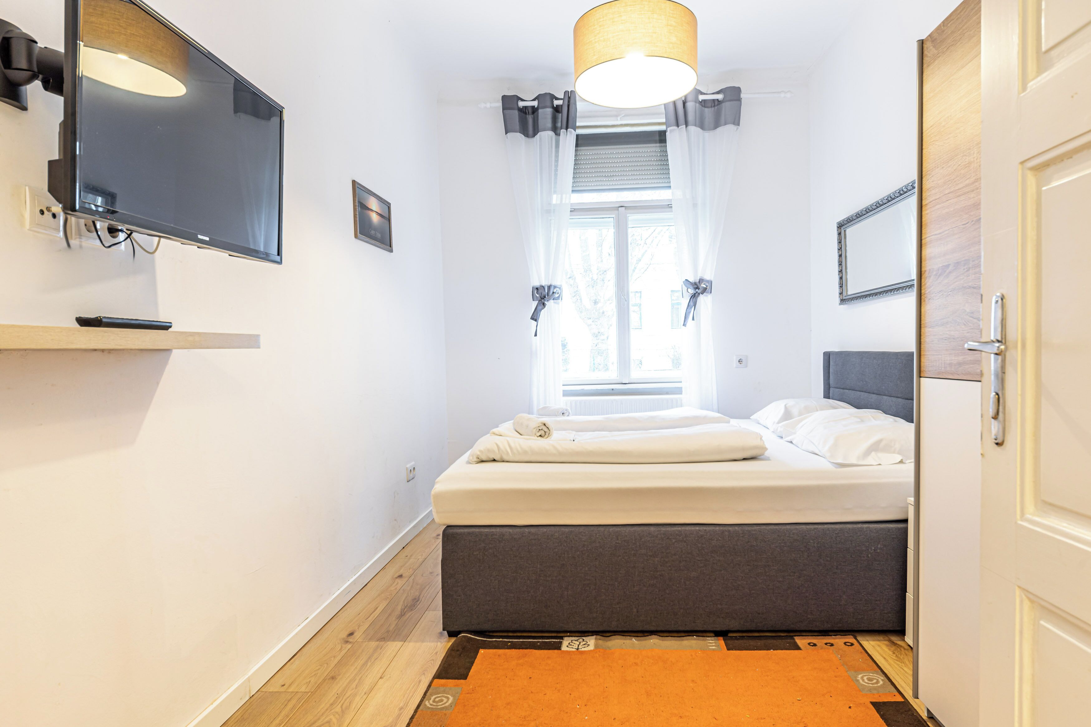 Une chambre à coucher avec un lit, un téléviseur fixé au mur, une fenêtre avec des rideaux et une porte.