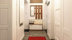 A narrow hallway with white walls, a wooden floor, a red rug, a mirror, and a small white cabinet.