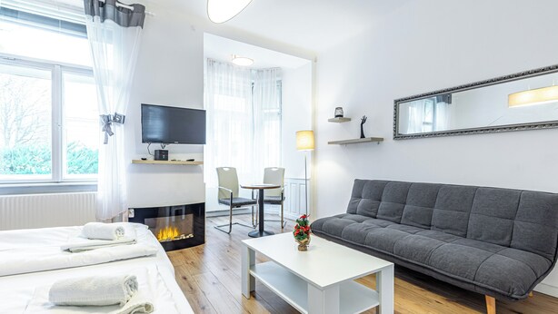 A modern hotel room with a sofa, a TV, a fireplace, a dining table, and a large window with curtains.