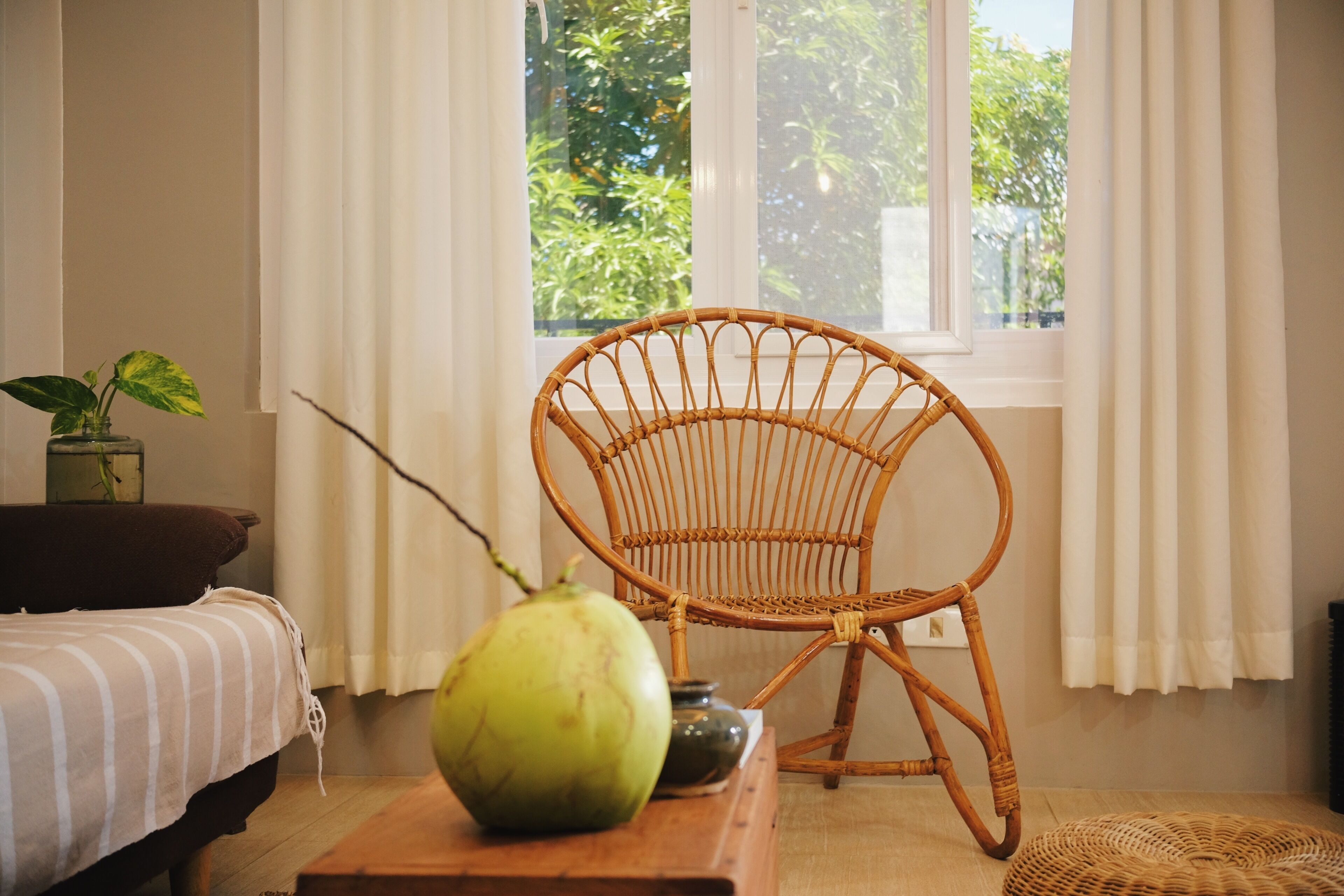 A room with a bed, a wicker chair, a table with a green object, and a window with curtains.