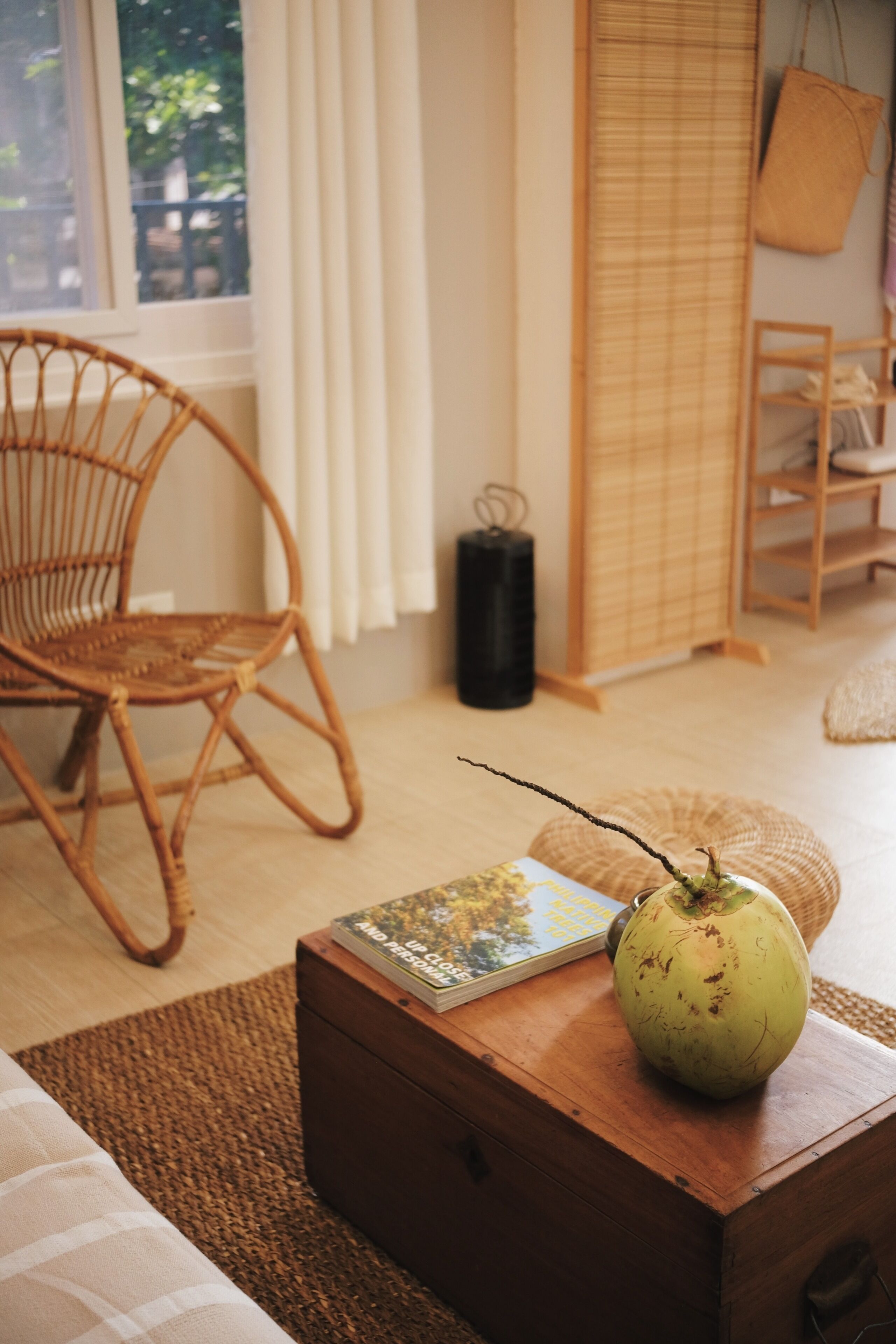 A room with a wicker chair, a wooden nightstand with a book and a green object, a woven mat, and a window with curtains.