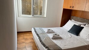 Ein ordentlich bezogenes Bett mit weißen Laken und einem dunklen Kopfkissen, ein Fenster mit Blick auf Bäume und Holzschränke über dem Bett.