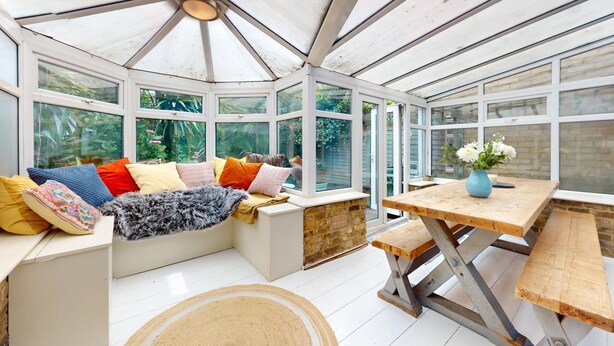 A conservatory with a glass ceiling, a seating area with colorful cushions, a wooden table with a vase of flowers, and a stone fireplace.
