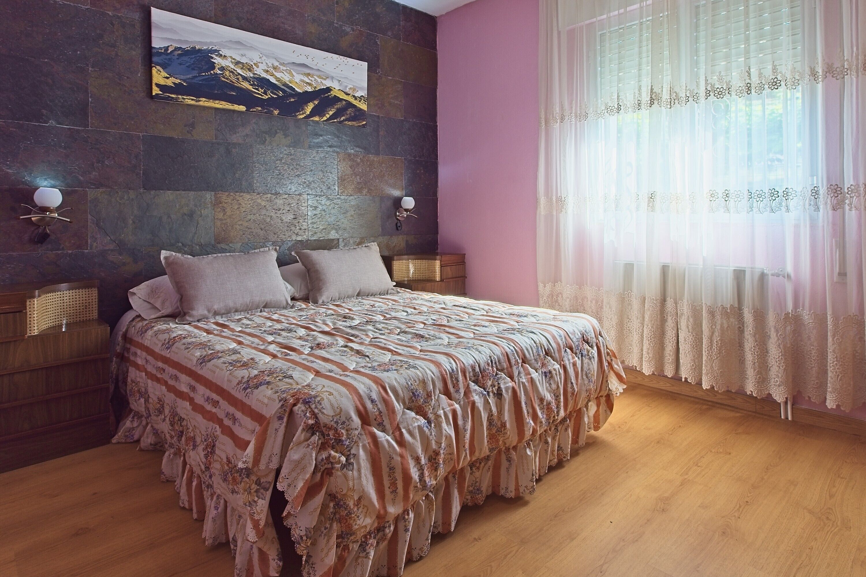 Une chambre avec un grand lit, un mur en pierre, un tableau représentant une montagne et une fenêtre ornée de rideaux légers.
