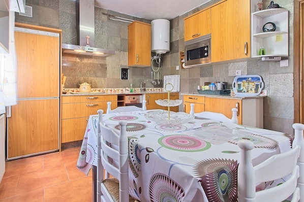 Une cuisine avec des meubles de rangement en bois, un four à micro-ondes, un chauffe-eau et une table à manger recouverte d’une nappe à motifs.