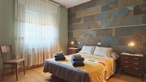 Un dormitorio con una cama, dos mesitas de noche, una silla y un ventanal con cortinas.