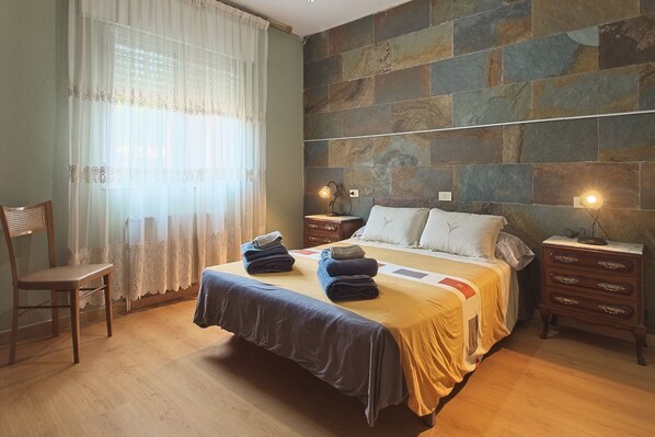 Un dormitorio con una cama, dos mesitas de noche, una silla y un ventanal con cortinas.