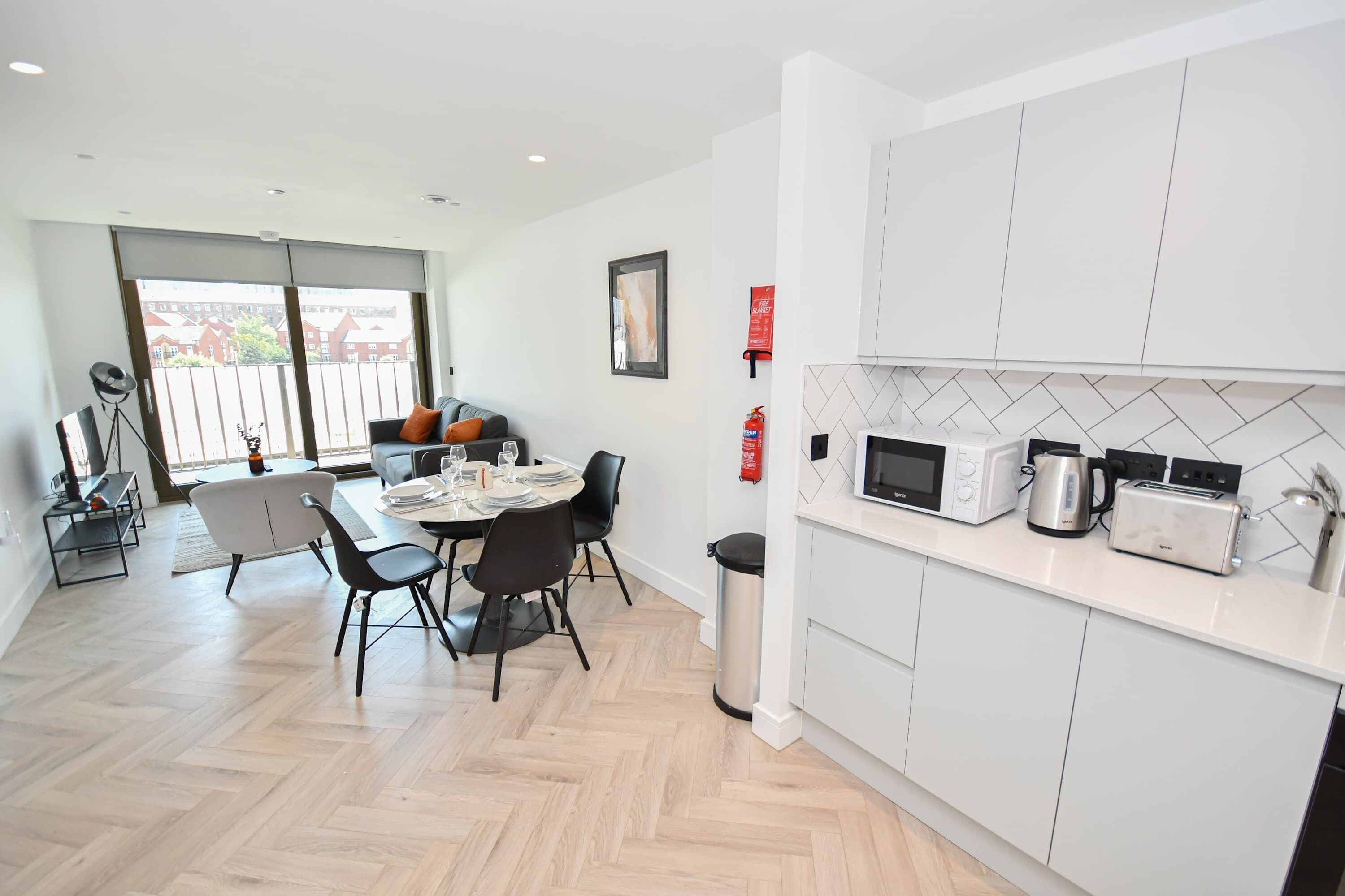Une cuisine moderne avec des meubles blancs, un four à micro-ondes, un grille-pain et une bouilloire. On y trouve également un coin repas avec une table et des chaises, ainsi qu’une grande fenêtre donnant sur des immeubles.