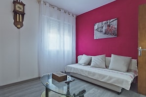 A room with a sofa, a glass coffee table, a clock, and a painting on the wall.