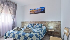 A bedroom with a checkered bedspread, a headboard with a stone pattern, a wall-mounted painting, and a small round table with a lamp.