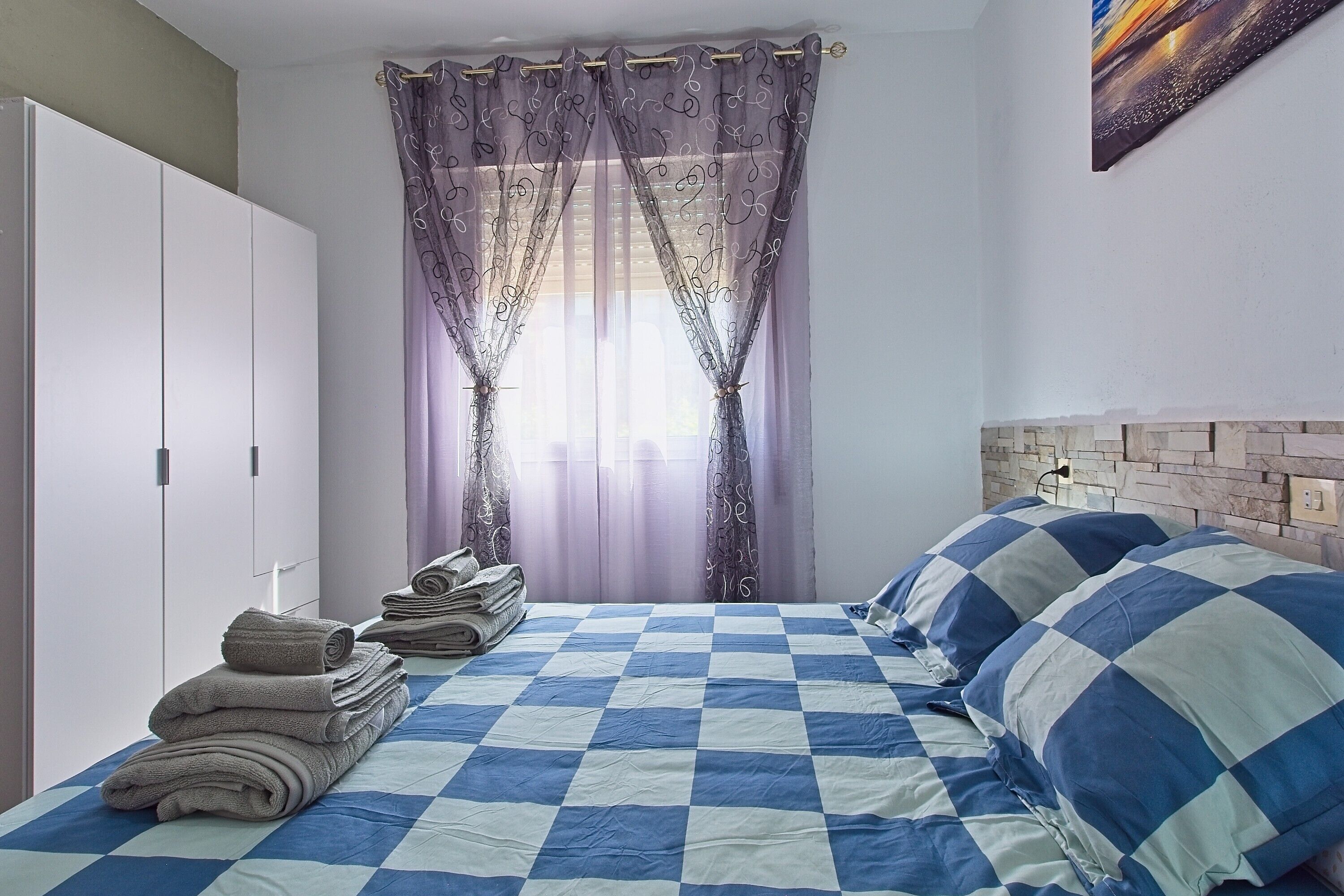 Une chambre avec une couvre-lit à carreaux, une pile de serviettes, une armoire et une fenêtre ornée de rideaux à motifs.