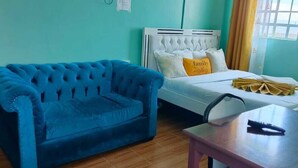 A room with a blue sofa, a white bed with a yellow pillow, and a small table with a remote control.