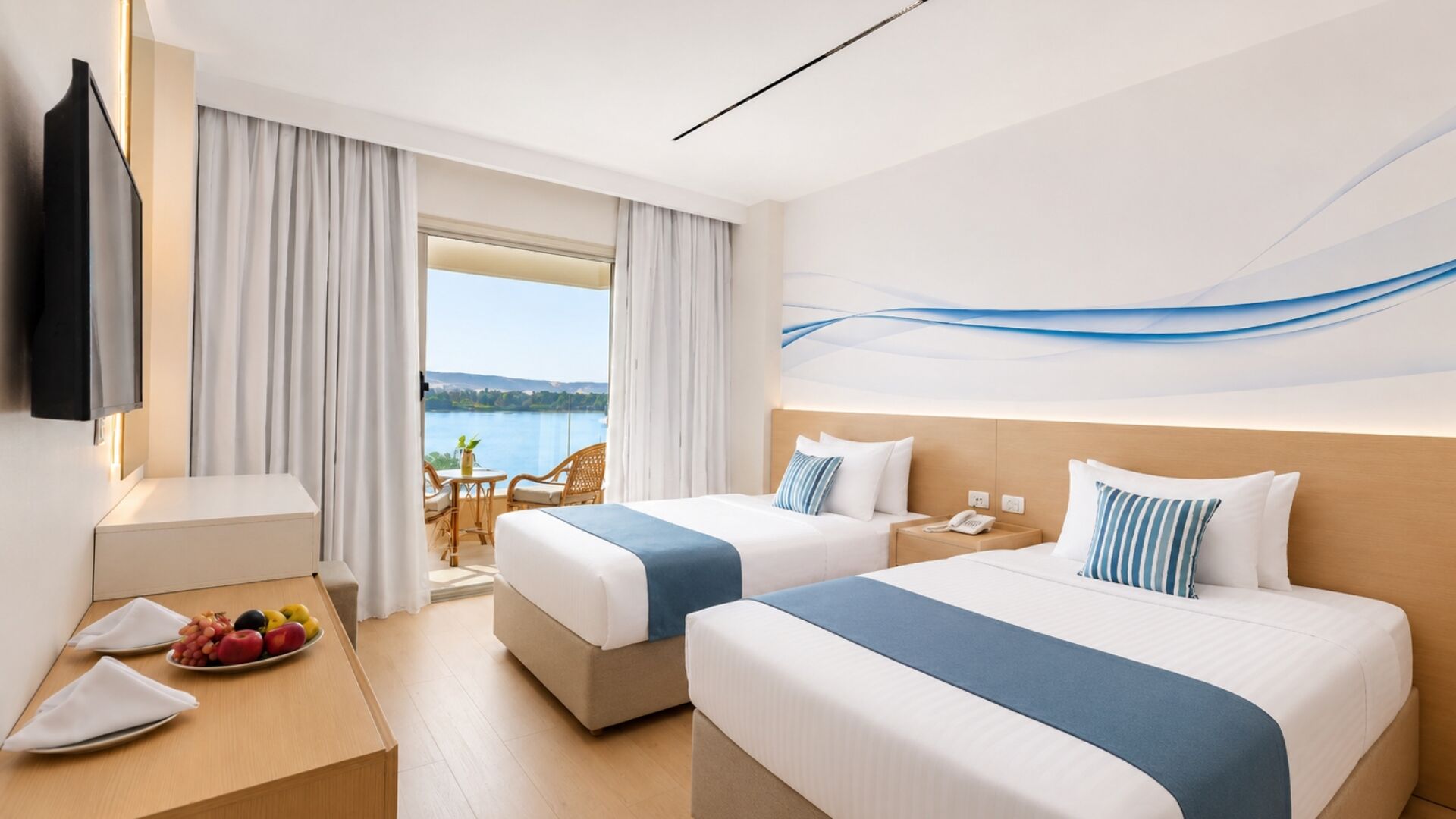 A hotel room with two beds, a desk with a fruit plate, a TV, and a view of a lake.