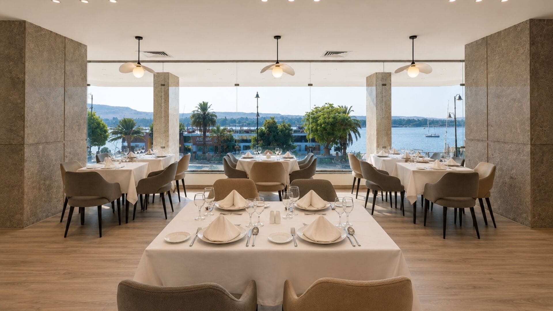 En restaurang med bord som är dukade för middag, stora fönster med utsikt över havet och kullarna samt hängande lampor.