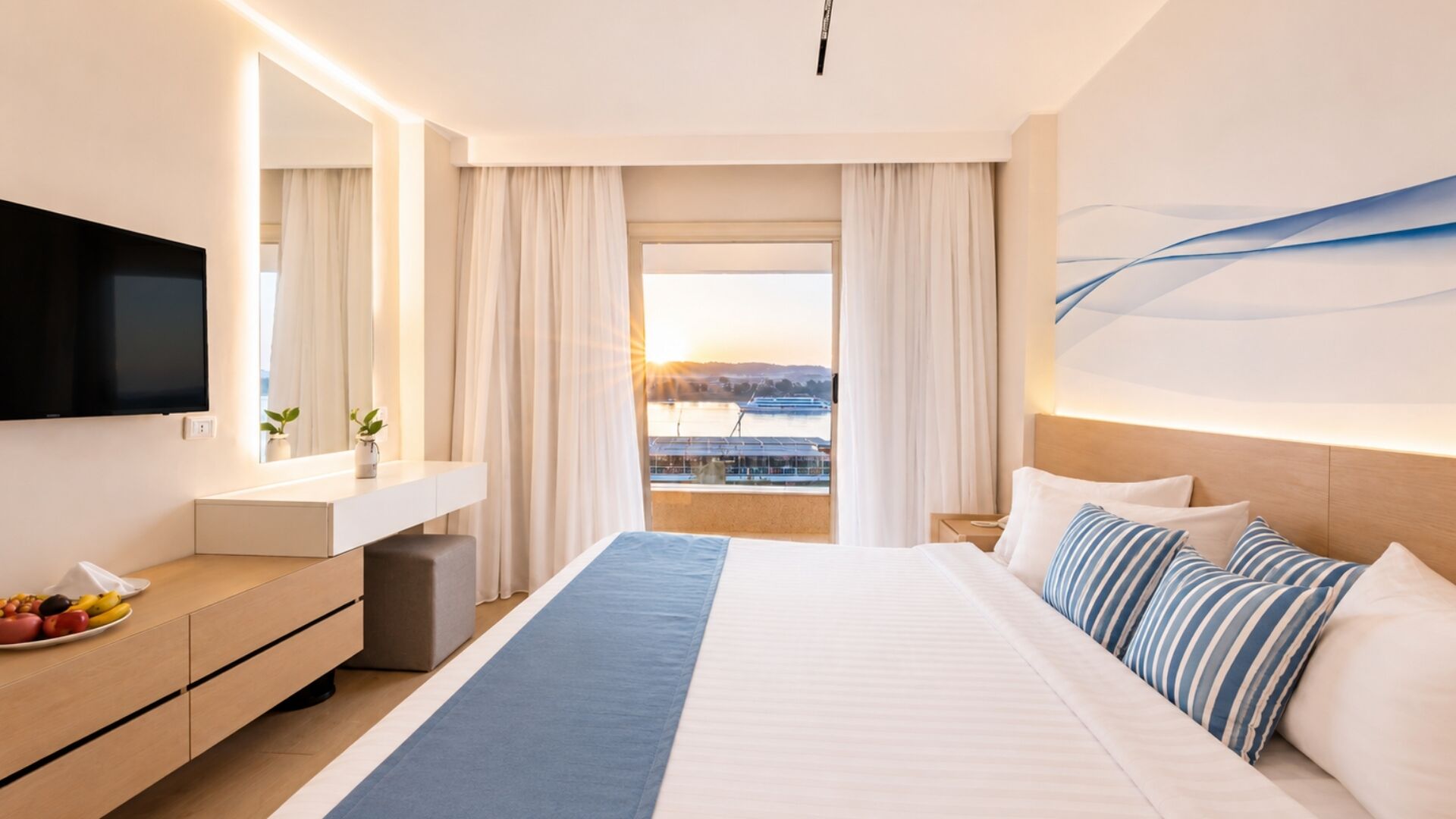 Ein Hotelzimmer mit Bett, Fernseher, Schreibtisch mit Pflanze und Blick auf ein Gewässer.