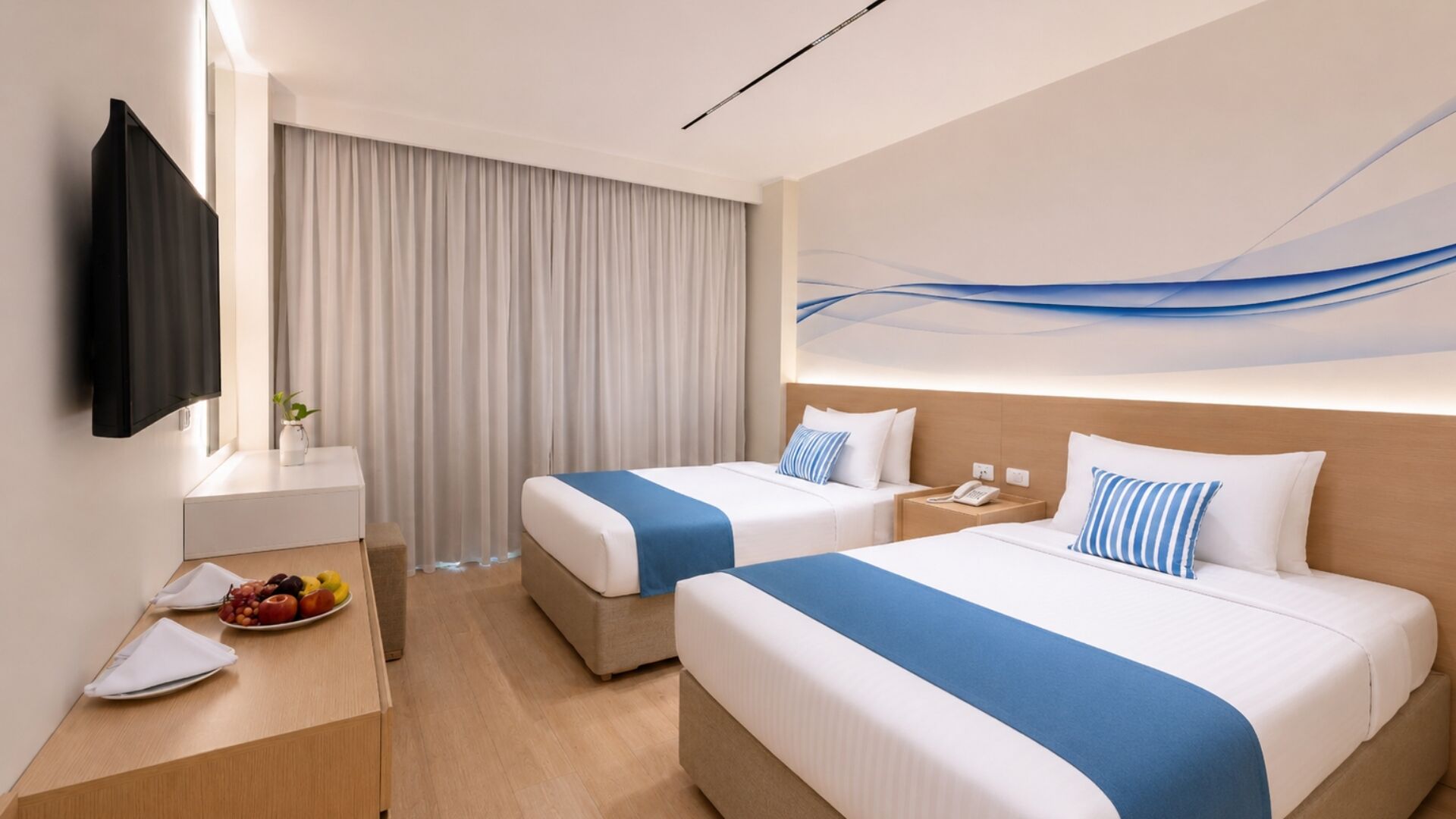 Une chambre d’hôtel avec deux lits, un bureau sur lequel se trouve une assiette de fruits, un téléviseur à écran plat et un tableau accroché au mur.