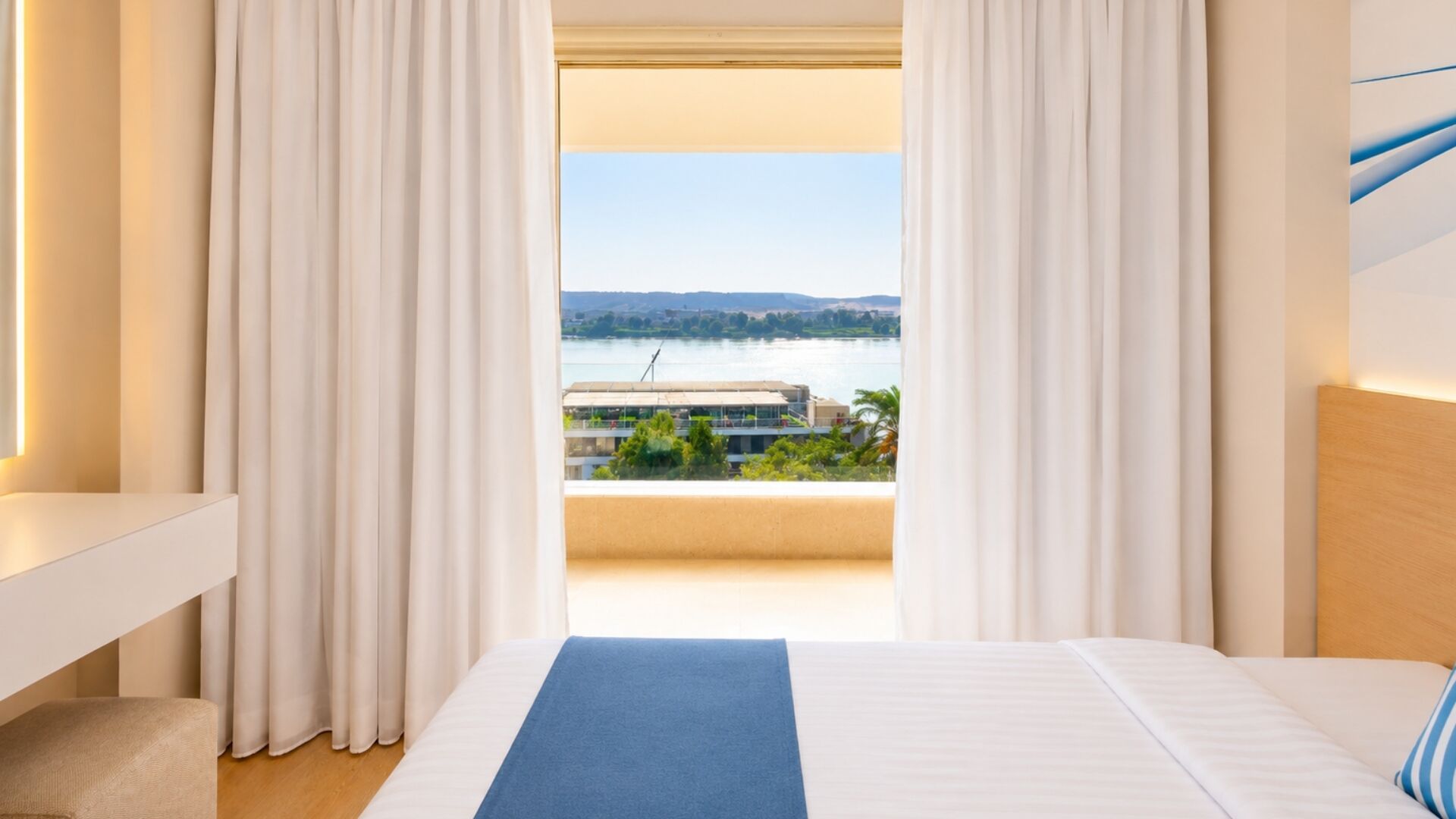 Ein Hotelzimmer mit Bett, weißen Vorhängen, Blick auf ein Gewässer und einem Balkon mit Geländer.