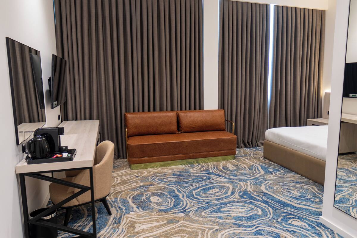 A hotel room with a brown sofa, a desk with a coffee maker, a bed, and a large window with curtains.