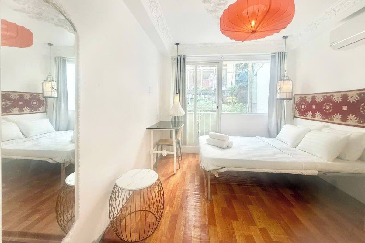 A hotel room with two beds, a large window, a mirror, a round table, and a decorative ceiling light.