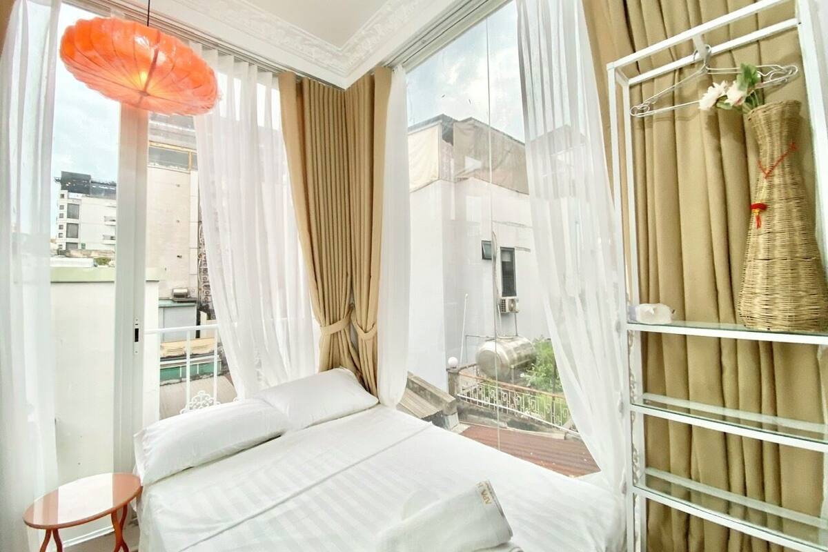 A hotel room with a bed, a small table, a hanging basket with flowers, and a view of the city through large windows.