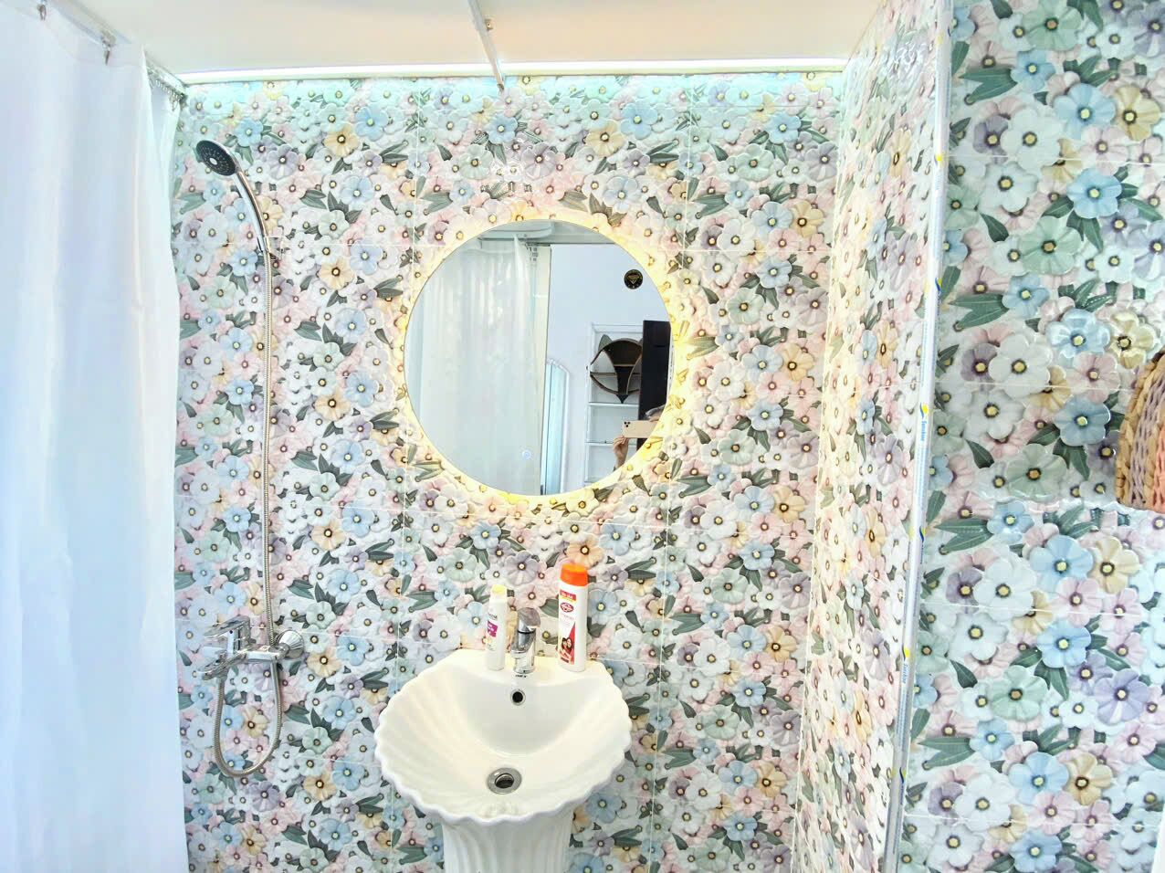 A bathroom with a floral wallpaper, a round mirror, and a sink with toiletries.
