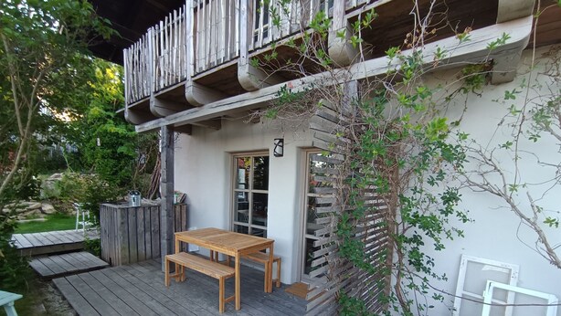 Une terrasse en bois avec une table et des bancs, entourée de verdure et bordée d’un bâtiment blanc doté d’un balcon.