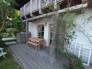 Una terraza de madera con una mesa y bancos, rodeada de follaje y un edificio blanco con balcón.