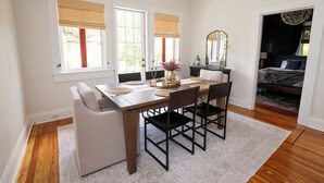 Un coin repas avec une table en bois, des chaises noires et un banc blanc. Il y a deux fenêtres avec des stores, un miroir et un lustre.