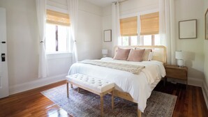 Une chambre à coucher avec un grand lit, une banquette, deux tables de chevet et une fenêtre avec des stores.