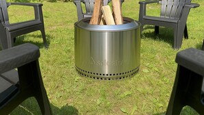 Un brasero de metal con dos sillas Adirondack a su alrededor, ubicado en un espacio al aire libre cubierto de césped y con árboles de fondo.