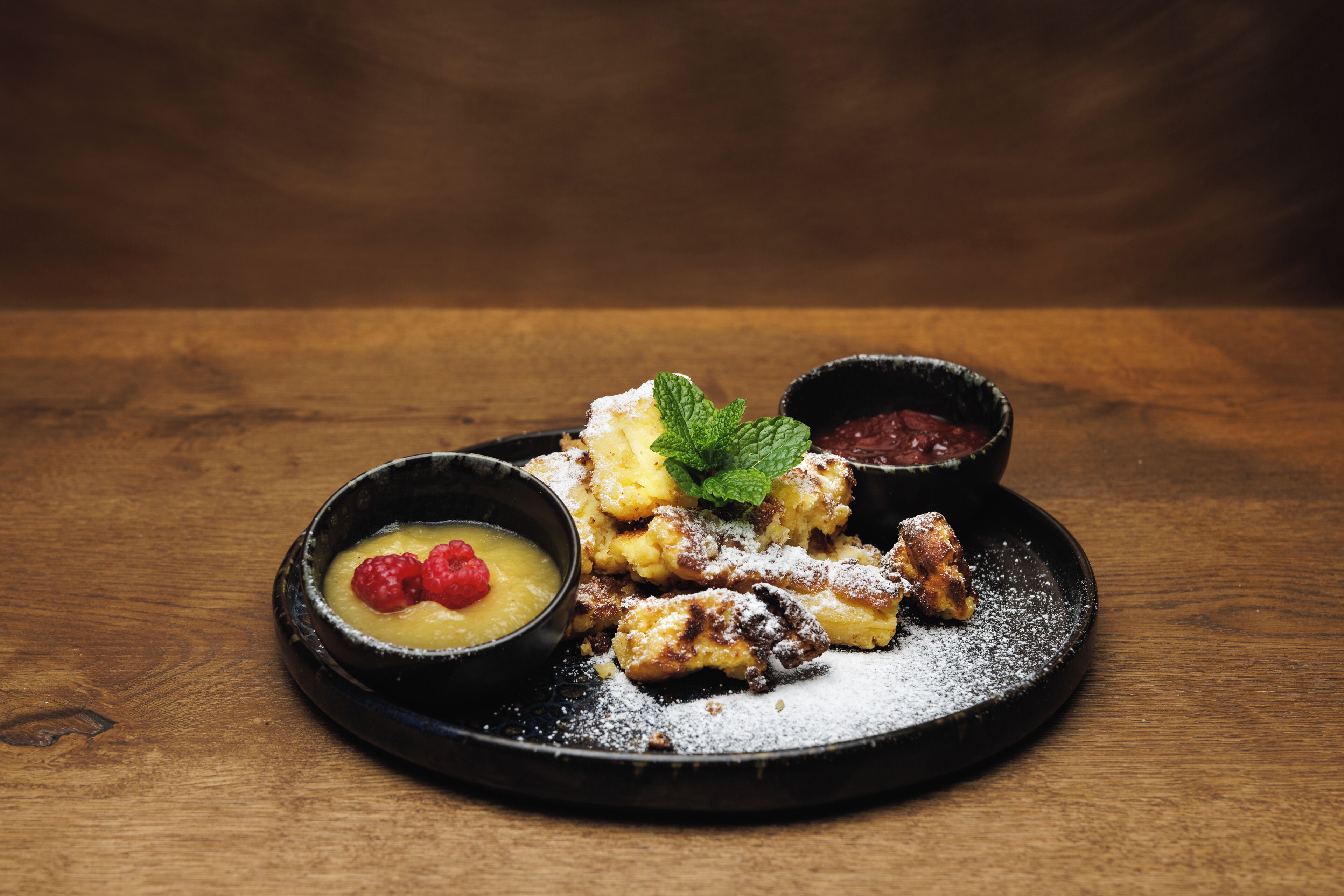 A dessert plate with powdered sugar, a mint leaf, and two small bowls of sauce.