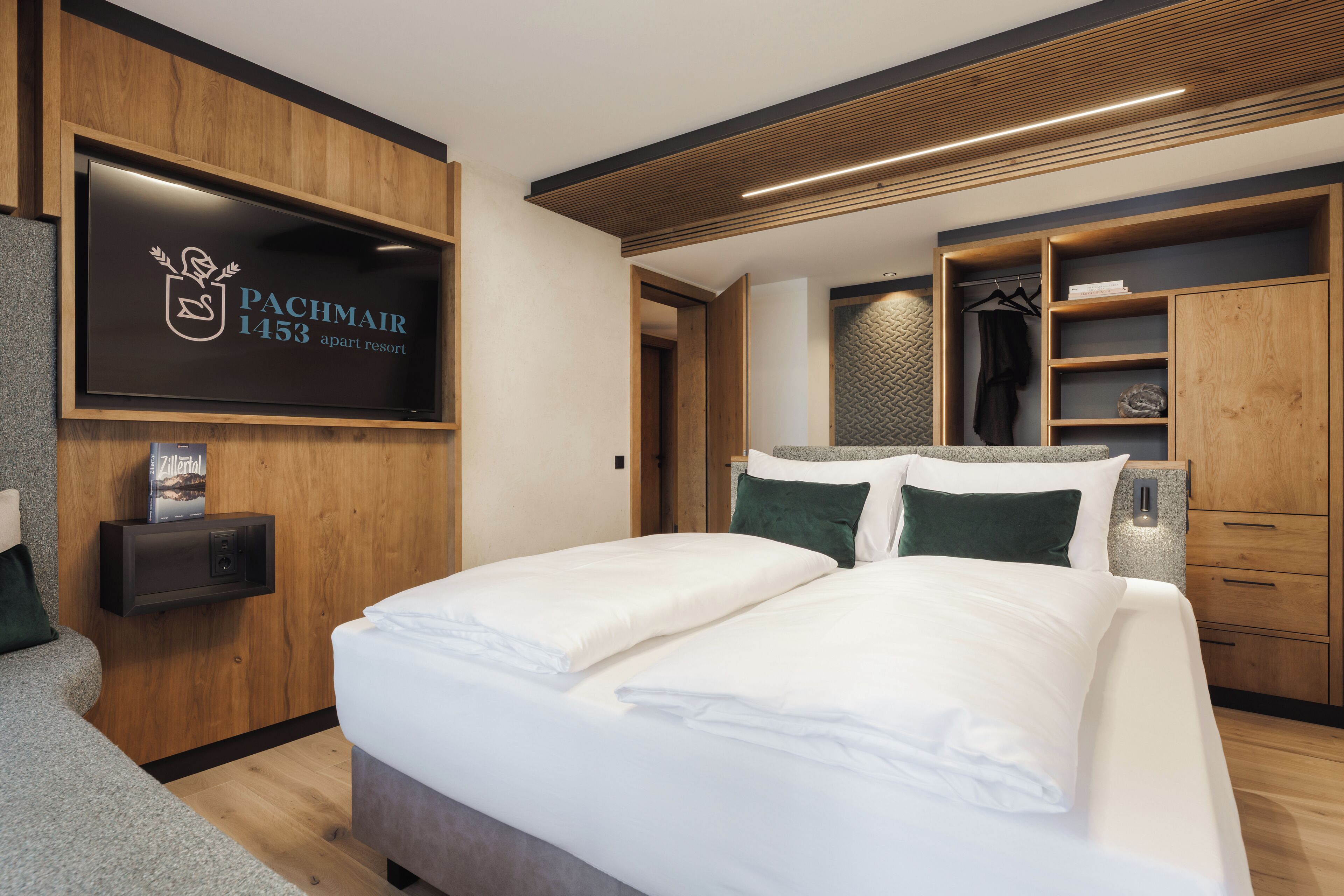 A modern bedroom with a large bed, built-in wardrobe, and a flat-screen TV displaying 'Pachmair 1453 apart resort'.
