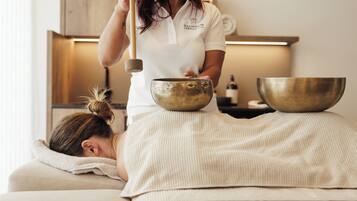 A person receiving a sound therapy session with two large bowls and a mallet in a room with a wooden cabinet and a lit lamp in the background.