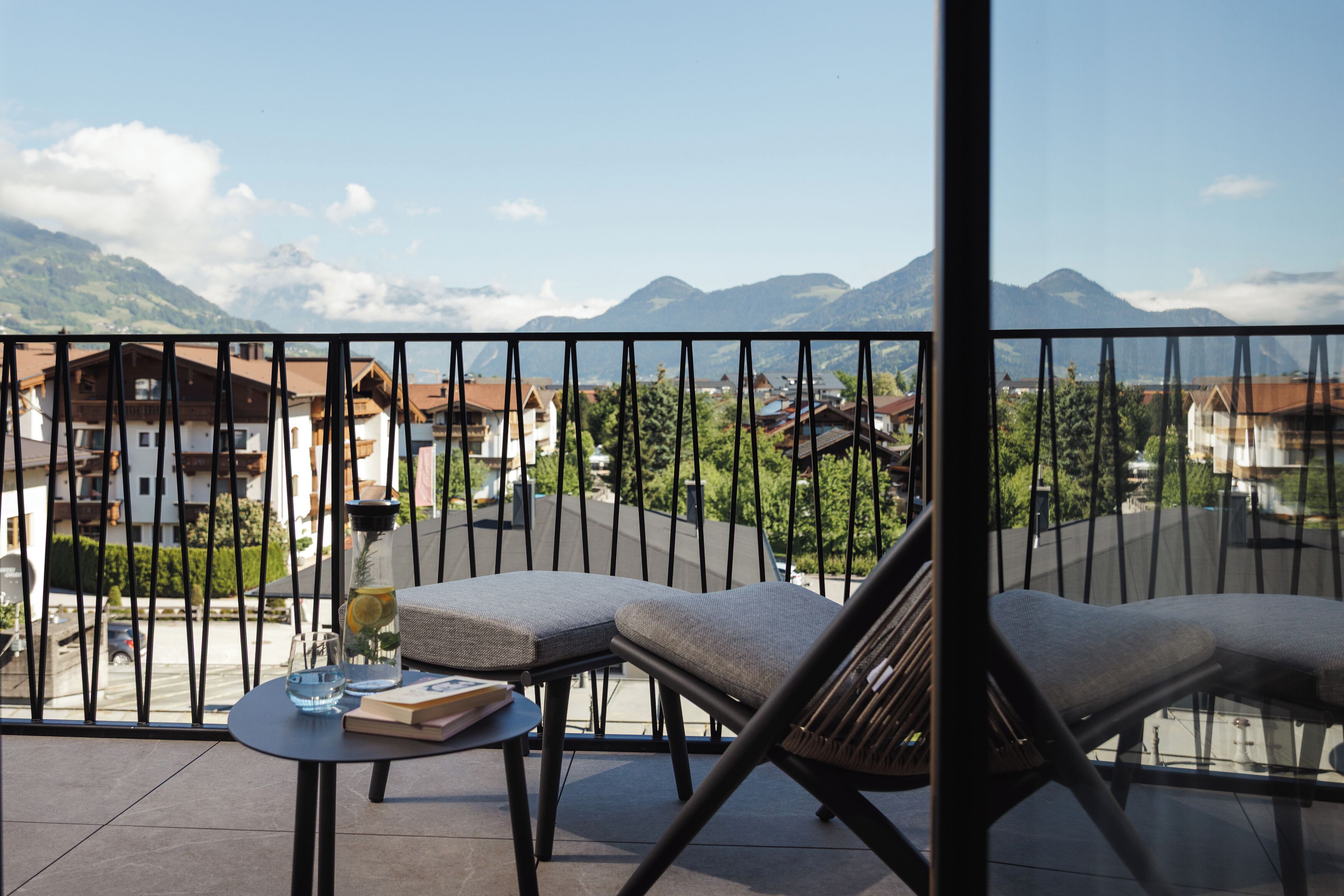 Un balcón con dos sillas acolchadas y una pequeña mesa, con vistas a un paisaje montañoso con casas.