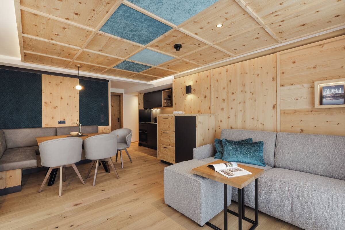 A modern interior with a wooden ceiling, a grey sofa, a small dining table with chairs, and a kitchen area.