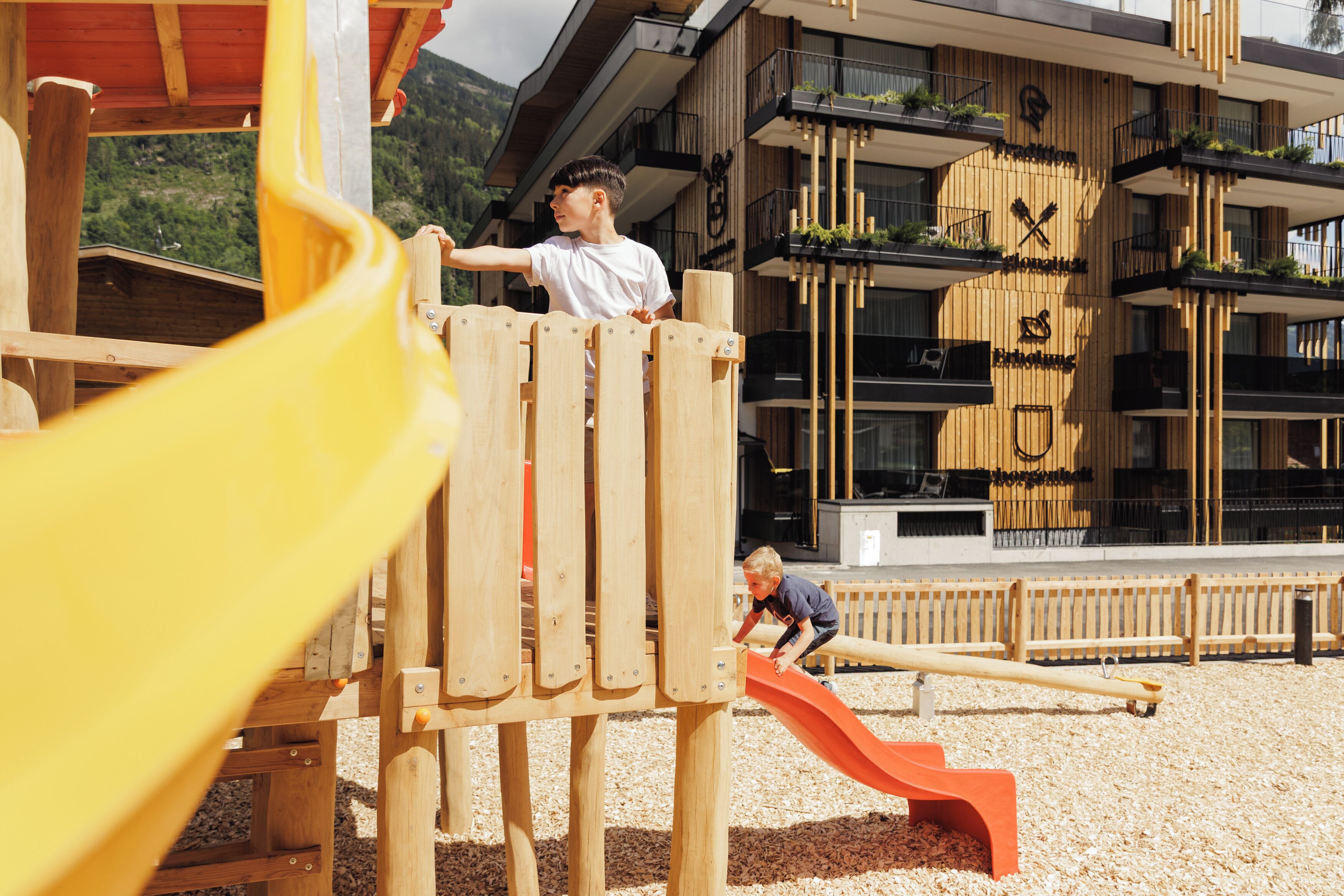 Un terrain de jeux doté de structures en bois et d’un toboggan, le tout situé devant un bâtiment moderne agrémenté de balcons et de plantes.