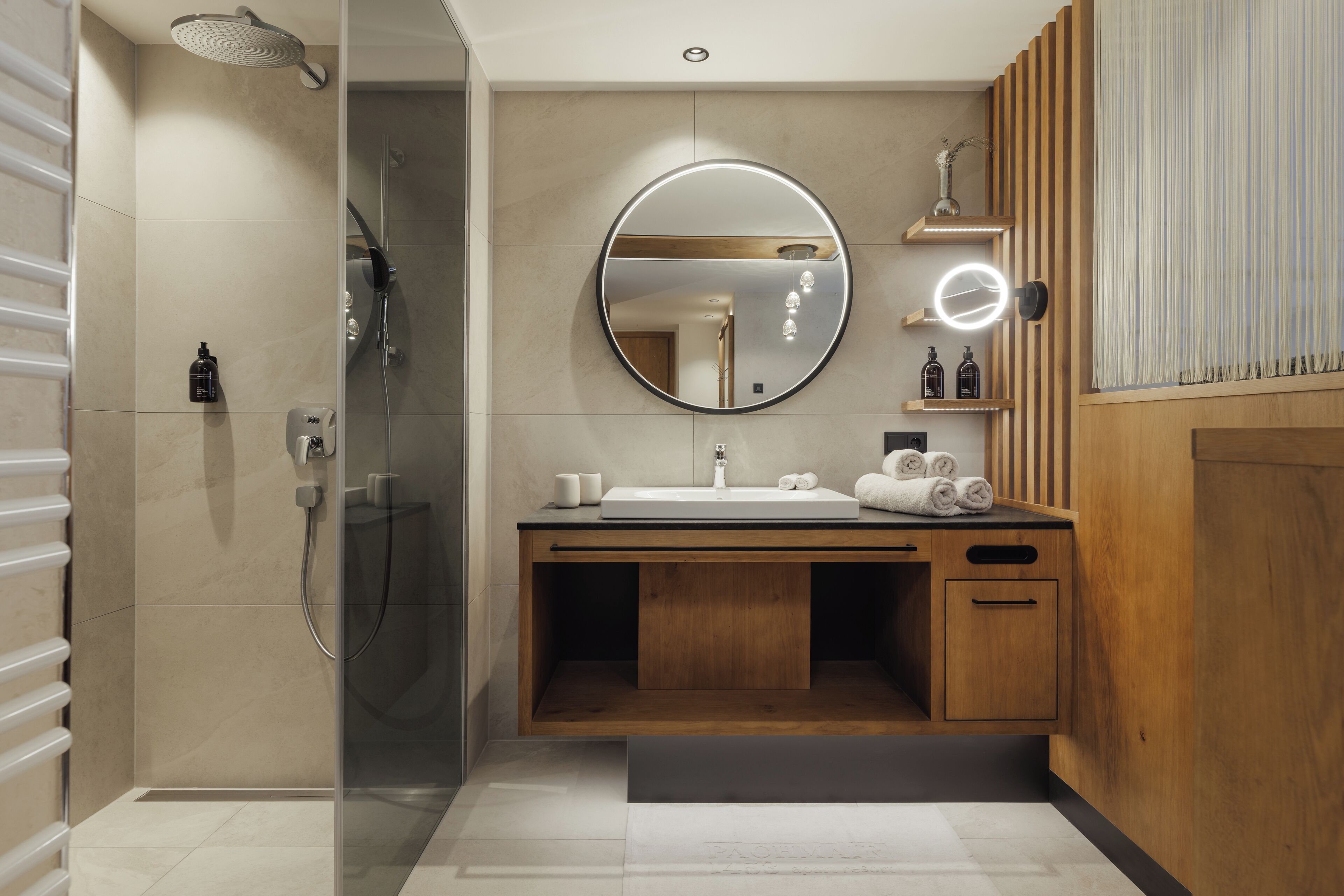 A modern bathroom with a glass-enclosed shower, a wooden vanity with a round mirror, and a built-in shelf with toiletries.