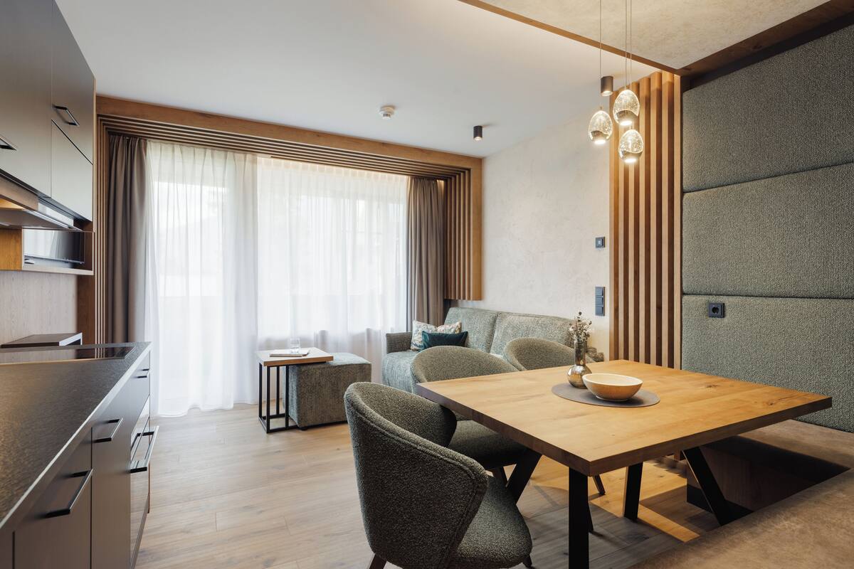 A modern kitchen with a wooden countertop and built-in appliances. There is a dining area with a wooden table and chairs. The room has large windows with curtains and a view of the outside.