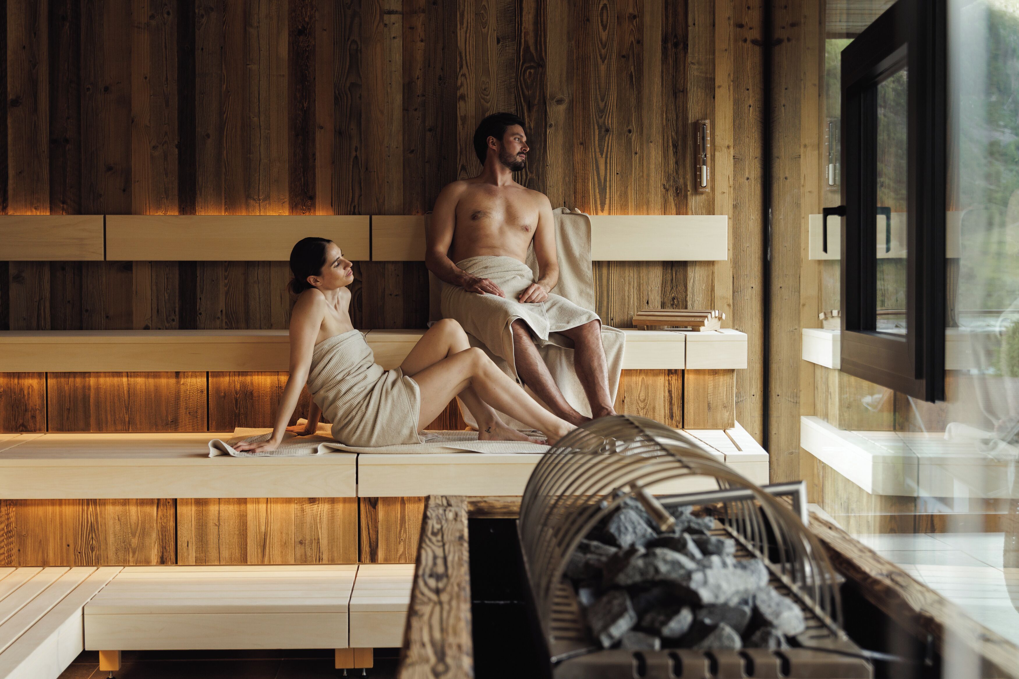 A wooden spa room with two people sitting on benches, a wooden wall, and a window.