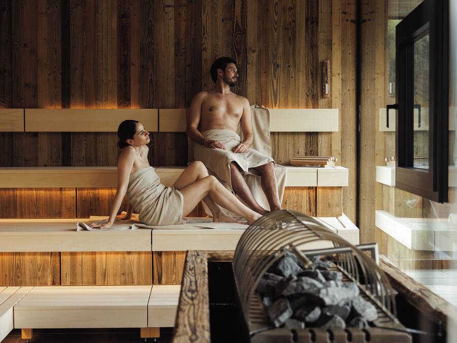 A wooden spa room with two people sitting on benches, a wooden wall, and a window.