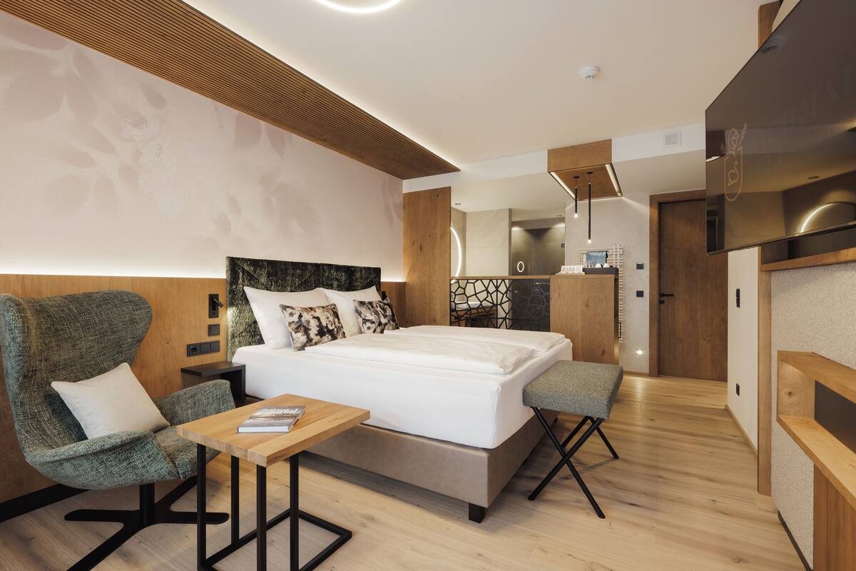 A modern hotel room with a large bed, a wooden headboard, a seating area with a table, and a small kitchenette.