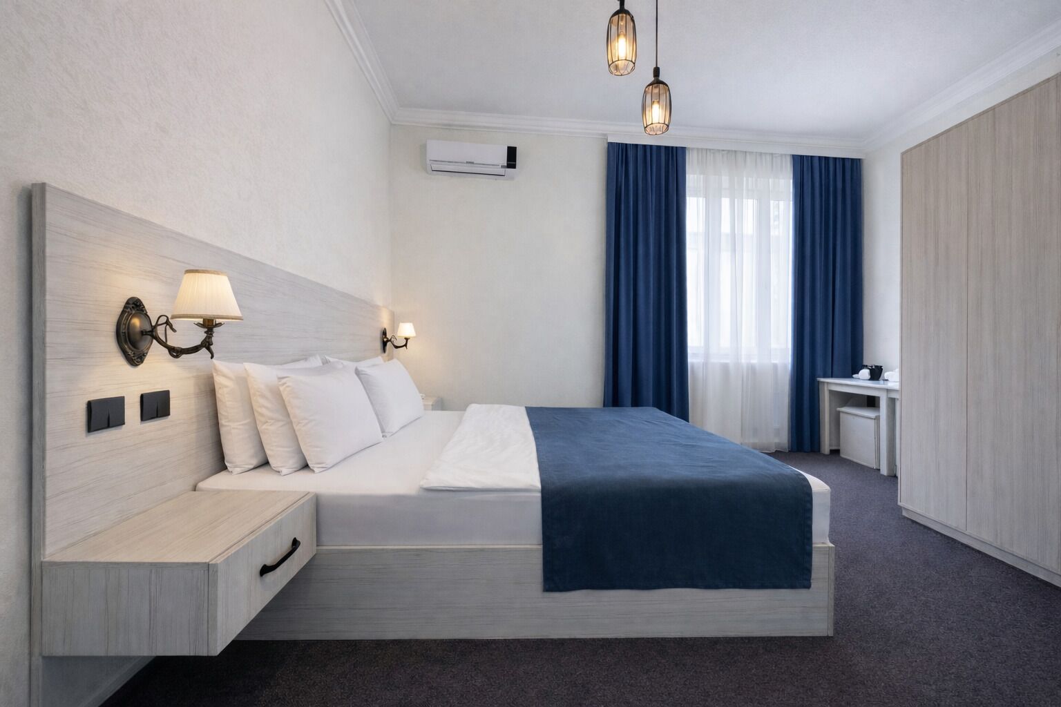 A hotel room with a large bed, bedside lamps, an air conditioning unit, a desk, and a wardrobe.
