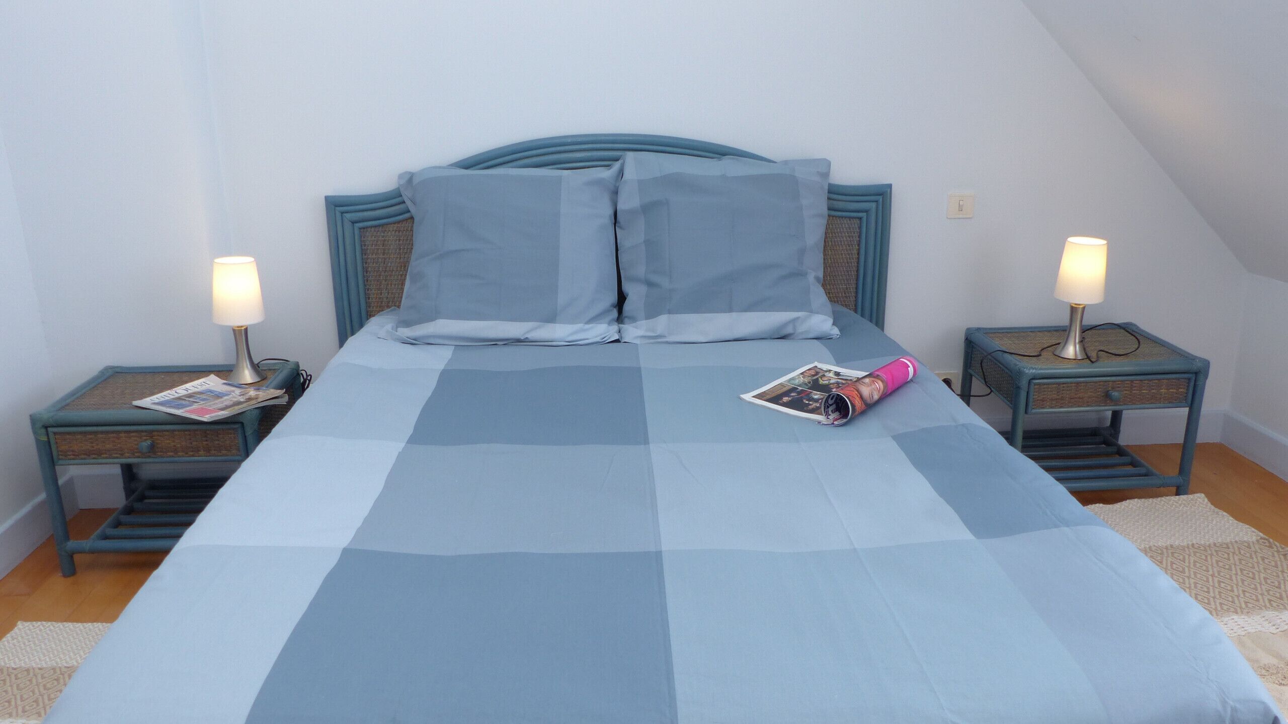 Ein ordentlich bezogenes Bett mit einer blau karierten Tagesdecke, zwei Nachttische mit Lampen und eine Zeitschrift auf dem Bett.