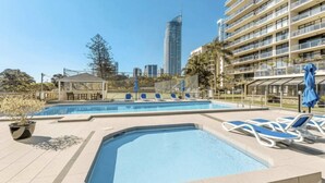 A pool area with lounge chairs and a gazebo, surrounded by a fence and palm trees, with a city skyline in the background.