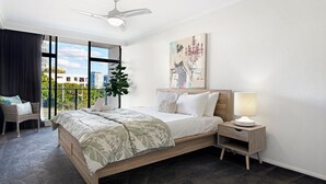 A modern bedroom with a large bed, a nightstand, a chair, a plant, and a view of the city through large windows.