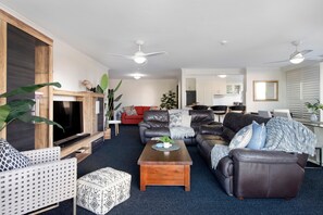 A modern living room with a flat-screen TV, leather sofas, a coffee table, and a dining area with a kitchen in the background. - Sunny Surfers Stay - Beach & Tram at your doorstep (Surfers Paradise)