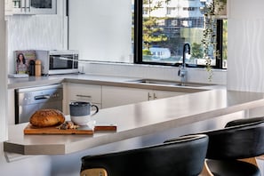 A modern kitchen with a large island, a built-in microwave, and a view of the city through the window. - Sunny Surfers Stay - Beach & Tram at your doorstep (Surfers Paradise)