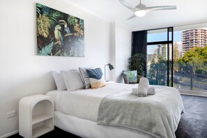 A modern bedroom with a large bed, a ceiling fan, a view of a cityscape through large windows, and a tropical-themed painting on the wall. - Sunny Surfers Stay - Beach & Tram at your doorstep (Surfers Paradise)